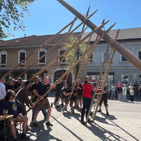 Eine Gruppe von Menschen hält große Holzstangen in einem Quadrat hoch, möglicherweise als traditioneller Tanz oder Veranstaltung.