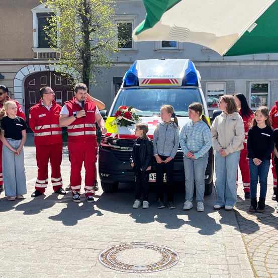 Ein Krankenwagen mit einer Blumendekoration auf der Motorhaube steht vor einer Gruppe von Menschen, darunter zwei Männer in roten Uniformen, die wahrscheinlich eine Rede halten.