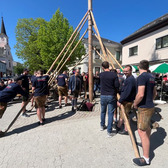 Eine Gruppe von Menschen in traditioneller bayerischer Kleidung hält große Stangen und nimmt an einem kulturellen Ereignis teil.