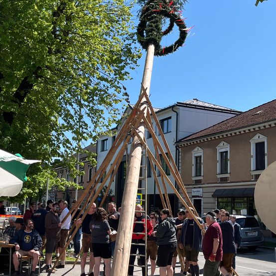 Eine hohe Holzkonstruktion mit einem Kranz oben steht in der Mitte der Straße. Mehrere Menschen stehen darum herum. Einige von ihnen halten Gläser. Ein Van ist am Straßenrand geparkt. Ein Baum und Gebäude sind im Hintergrund.