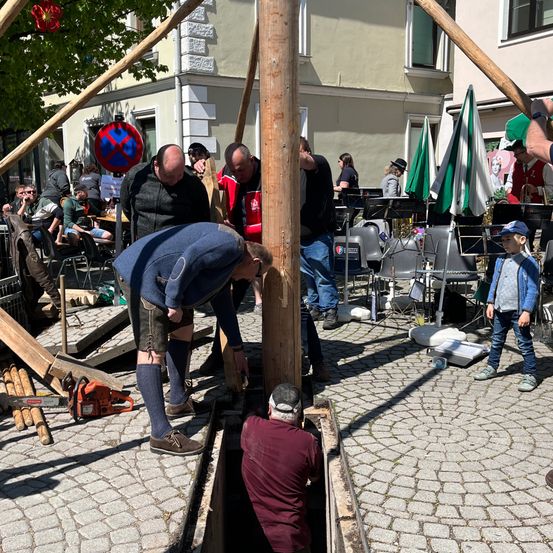 Männer und ein Junge arbeiten an einem Holzpfahl auf einem Kopfsteinpflasterplatz. In der Nähe stehen Stühle, Tische und Regenschirme.