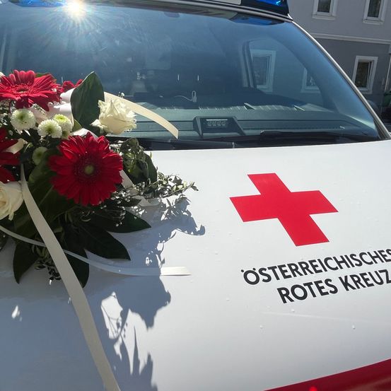Ein Krankenwagen mit einem Blumenstrauß vorne, der das Logo des Österreichischen Roten Kreuzes auf der Seite zeigt.