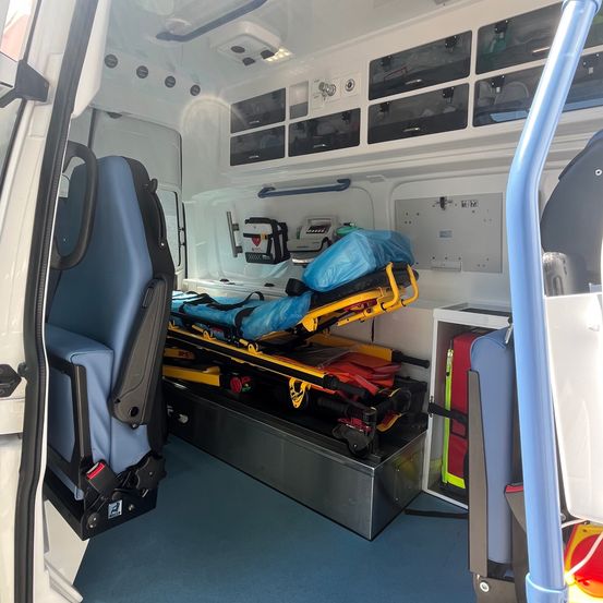 Das Innere eines Krankenwagens mit einer Trage und medizinischen Geräten in der Mitte, einem blauen Fahrersitz links und einer grauen Metall-Aufbewahrungsbox mit medizinischer Ausrüstung rechts.