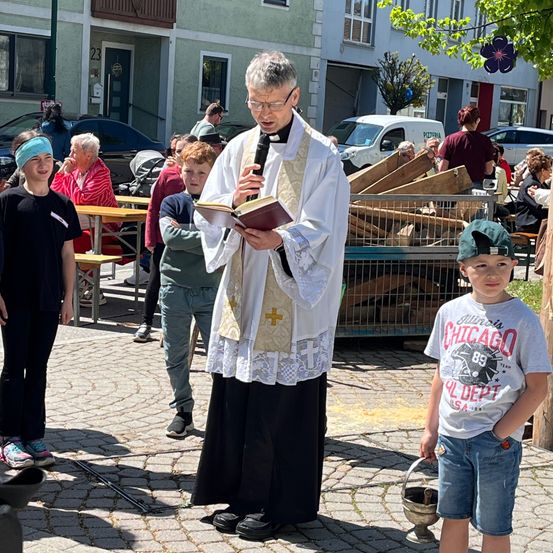 Ein Priester hält ein Buch und ein Mikrofon und spricht zu einer Gruppe von Kindern und Erwachsenen im Freien.