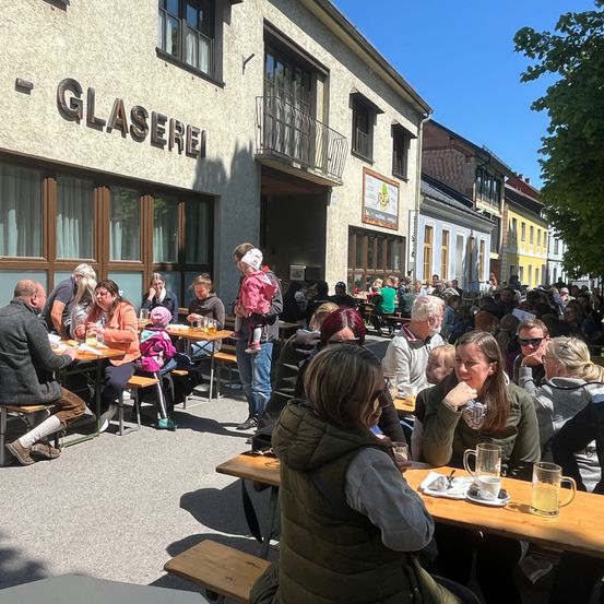 Eine Gruppe von Menschen sitzt draußen in einem Biergarten. Sie sitzen an Holztischen mit Tassen und Tellern darauf.