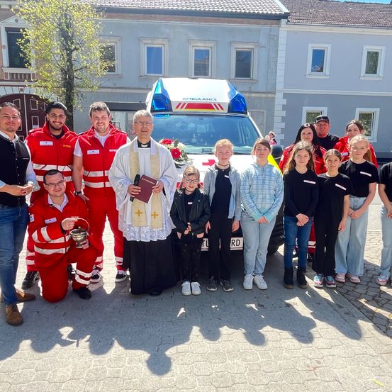 Eine Gruppe von Menschen in roten und schwarzen Uniformen steht vor einem Krankenwagen. Ein Priester, der ein Buch und eine Blume hält, steht neben einem Mädchen mit Brille.