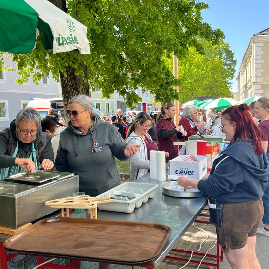 Mehrere Frauen bereiten Essen an einem Tisch im Freien unter einem grünen und weißen Sonnenschirm vor. Sie sind von anderen Menschen umgeben, und einige tragen eine Brille.