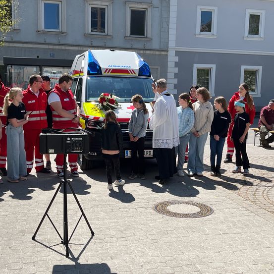 Ein Priester steht vor einem Krankenwagen mit einem Blumenstrauß. Eine Gruppe von Menschen steht um den Krankenwagen herum, einige in roten und weißen Uniformen.