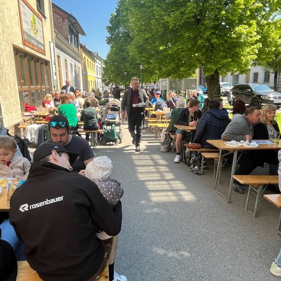 Menschen sitzen an Tischen im Freien an einem sonnigen Tag, ein Mann geht in der Mitte und Bäume und Gebäude im Hintergrund.