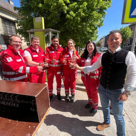 Eine Gruppe von Freiwilligen, die rote Uniformen tragen und Weingläser halten, steht unter einem Baum im Freien.