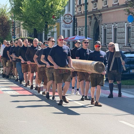 Eine Gruppe von Männern in traditioneller deutscher Kleidung trägt einen großen Baumstamm, während sie auf einer Stadtstraße gehen.