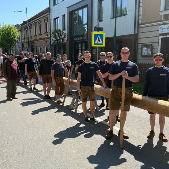 Eine Gruppe von Menschen in traditioneller bayerischer Kleidung, die Holzstäbe halten, steht in einer Reihe auf der Straße, mit Gebäuden und Straßenschildern im Hintergrund.