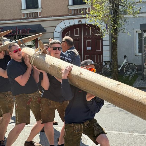 Fünf Männer in traditioneller Kleidung tragen einen langen Holzstab auf einer Straße. Sie tragen Sonnenbrillen und Mützen. Hinter ihnen steht ein Gebäude mit einer braunen Tür und einem Baum.