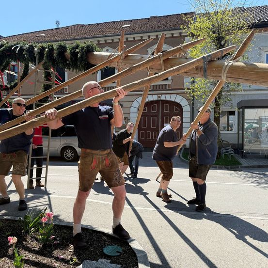 Fünf Männer in traditioneller bayerischer Kleidung halten große Holzspeere vor einem Gebäude mit einer braunen Tür.