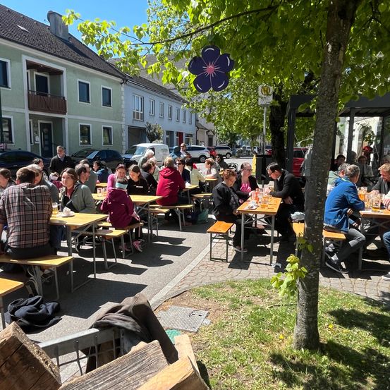 Eine Gruppe von Menschen sitzt an Holztischen unter einem Baum in einem Restaurant an der Straße. Im Hintergrund befinden sich Gebäude und geparkte Autos. Einige der Menschen tragen Brillen, Hüte und Sneakers. Es gibt Jacken, Tassen und andere Gegenstände auf den Tischen.