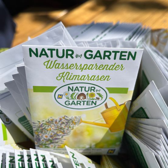 Ein Stapel weißer Samenpakete mit der Aufschrift 'Natur im Garten' auf einer Holzoberfläche, mit einem gelben Gießkannen- und Igellogo.