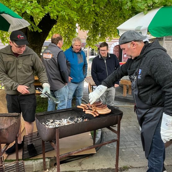 Fünf Männer stehen um einen Grill herum. Ein Mann grillt Würste auf dem Grill, ein anderer hält eine Zange und die anderen schauen zu.