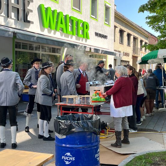 Eine Gruppe von Menschen in traditioneller deutscher Kleidung wartet in einer Schlange vor einem Restaurant namens Walter. Eine Frau serviert Essen an einem Tisch mit einem blauen Fass in der Nähe.