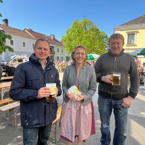 Drei Menschen stehen draußen, halten Getränke in den Händen und lächeln. Sie sind in der Nähe von Tischen und einem Krankenwagen. Dahinter befinden sich Häuser und Bäume.