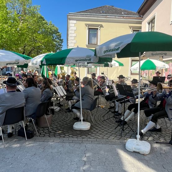 Eine Brassband spielt auf einer Kopfsteinpflasterstraße, wobei eine Gruppe von Musikern Trompeten und Posaunen spielt. Um sie herum sind Schirme für Schatten aufgestellt. Im Hintergrund befinden sich Gebäude, Autos und Bäume.