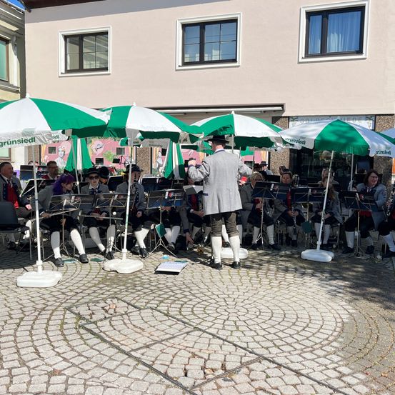 Eine Musikband spielt im Freien mit Schirmen vor einem Gebäude. Die Musiker tragen formelle Kleidung.