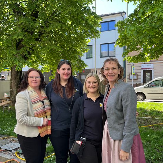 Vier Frauen stehen lächelnd vor einem Baum, mit einem weißen Gebäude und einem geparkten Auto im Hintergrund.