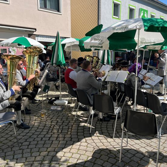 Ein Blasorchester spielt auf einem gepflasterten Außenbereich, mehrere Personen sitzen auf Stühlen.