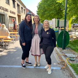 Drei Frauen stehen vor einem Gebäude. Sie lächeln und posieren für ein Foto. Hinter ihnen sind Tische und Bänke und ein grünes Objekt auf dem Rasen. Das Gebäude hat die Wörter Schleierei und Glaserei darauf. Bäume und Pflanzen sind in der Gegend. In der Ferne sitzen Menschen auf den Bänken.