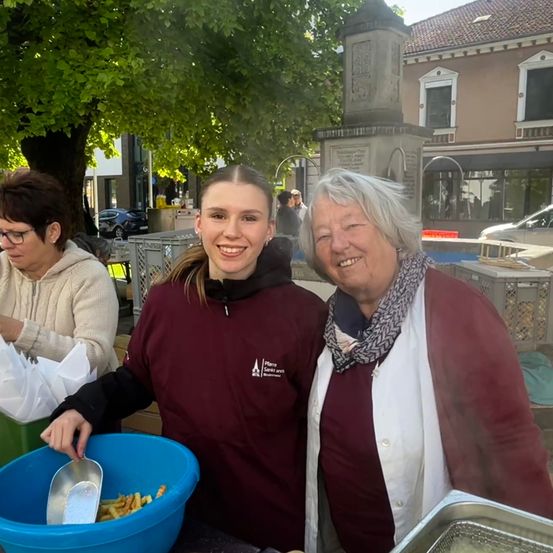 Zwei Frauen stehen vor einem Tisch im Freien. Eine Frau trägt ein kastanienbraunes Shirt und lächelt, während sie einen Schöpflöffel in einer blauen Schüssel hält. Die andere Frau trägt ein weißes Hemd und einen Schal und lächelt in die Kamera.