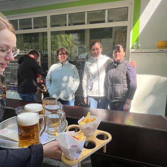 Eine Frau steht hinter einer Bartheke und hält ein Tablett mit Bier und Pommes, während drei Frauen hinter ihr zusehen.