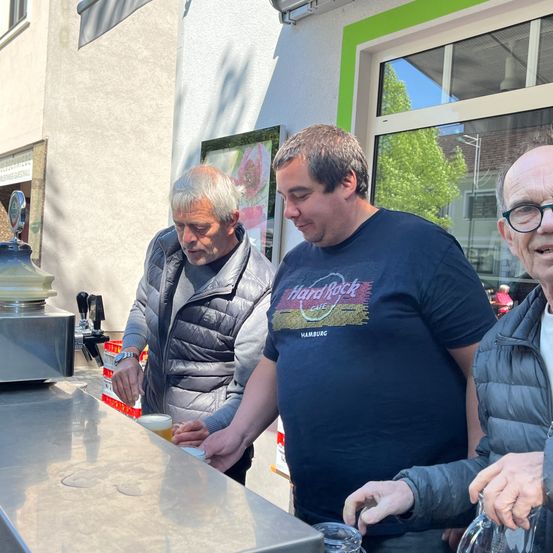 Drei Männer stehen an einer Bartheke, einer von ihnen trägt ein schwarzes T-Shirt mit dem Hard Rock Cafe Logo, und sie scheinen zu reden.