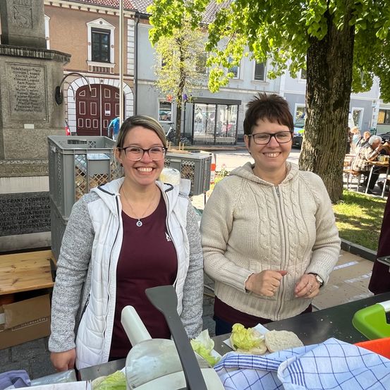Zwei Frauen stehen vor einem Tisch mit Salat. Eine Frau trägt eine Halskette und eine Brille, die andere eine Uhr. Hinter ihnen steht ein Mülleimer und ein Baum. In der Ferne befindet sich ein Gebäude mit Glastüren und Fenstern.