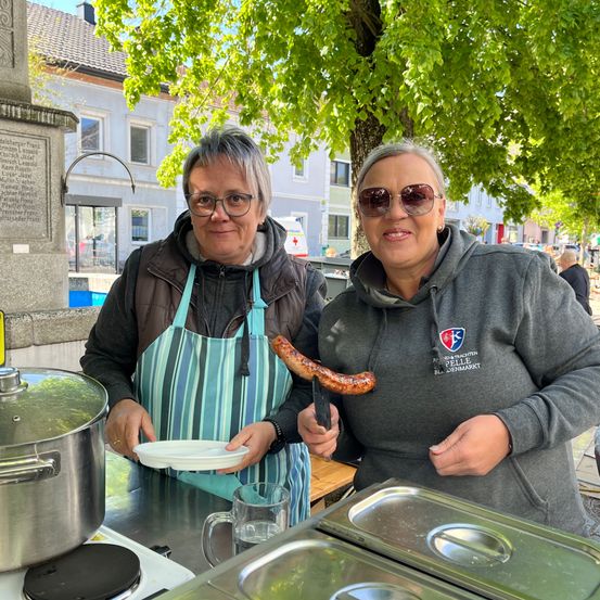 Zwei Frauen kochen draußen auf einem Markt, eine trägt eine blaue Schürze und hält eine Wurst, die andere trägt ein graues Sweatshirt mit Logo auf der Vorderseite.