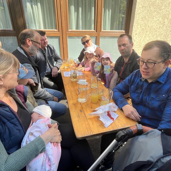 Eine Gruppe von Menschen sitzt um einen Tisch herum vor einem Gebäude. Sie lächeln und scheinen eine gute Zeit zu haben. Einige halten Getränke in Gläsern und eine Person hält ein Baby.