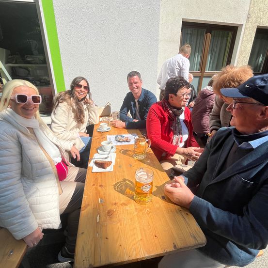 Eine Gruppe von Menschen sitzt draußen an einem Tisch, lächelt und schaut zur Kamera. Eine Frau links trägt eine Sonnenbrille und eine weiße Jacke. Vor ihnen stehen Tassen und Teller mit Essen. Ein Mann rechts trägt einen Hut und eine Brille. Hinter ihnen befindet sich ein Gebäude mit einer Glastür.