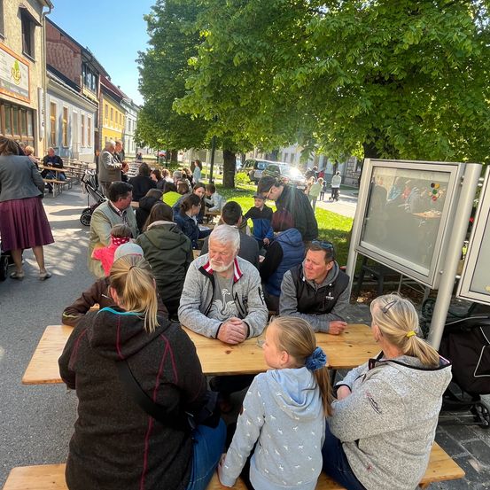 Eine Gruppe von Menschen sitzt an einem Tisch in einem Park an einem sonnigen Tag. Einige tragen Jacken, andere nicht. Ein Kinderwagen steht in der Nähe.