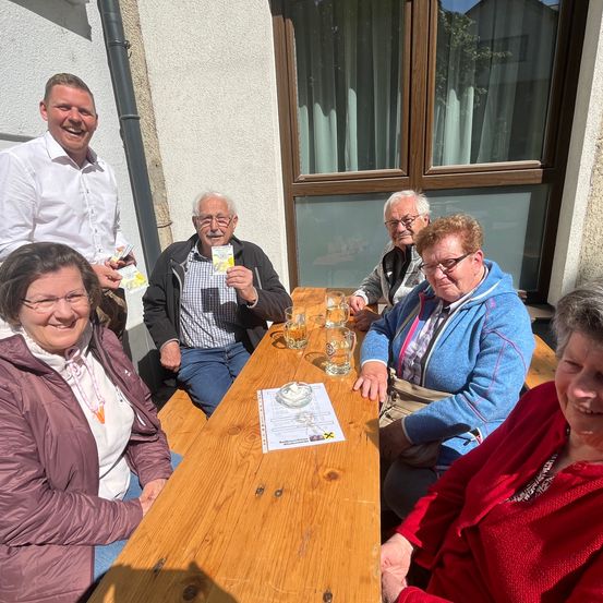 Eine Gruppe von Menschen sitzt draußen an einem Tisch, wobei zwei Männer Gläser mit Bier halten und eine Frau ein gelbes Ticket in der Hand hat.