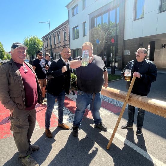 Fünf Männer stehen auf einer Straße, von denen einer ein Glas Bier in der Hand hält. Vor ihnen steht eine hölzerne Barrikade.