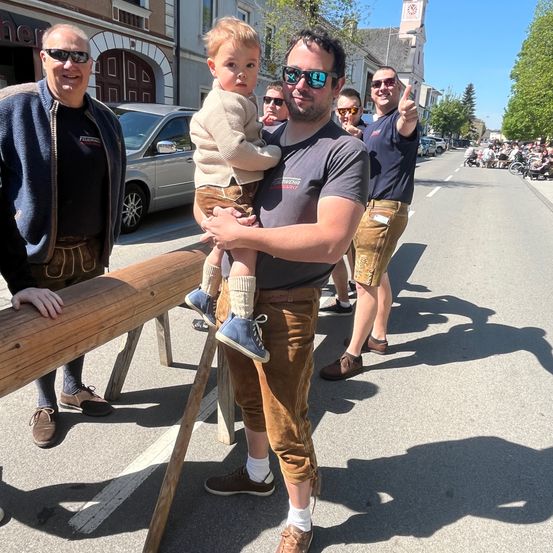 Drei Männer stehen in traditioneller Kleidung am Straßenrand. Der Mann in der Mitte hält einen kleinen Jungen. Eine Person steht auf einer hölzernen Barrikade. In der Ferne befinden sich geparkte Autos, Gebäude und Bäume.