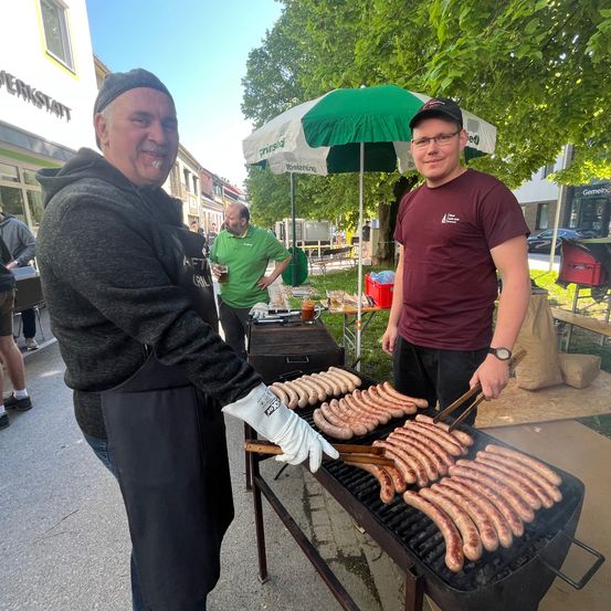 Zwei Männer grillen Würste im Freien. Ein Mann trägt einen weißen Handschuh und hält eine Zange. Der andere Mann trägt eine Uhr und hält ein Glas Bier. Im Hintergrund steht ein grüner Schirm, ein Tisch und ein Mülleimer.