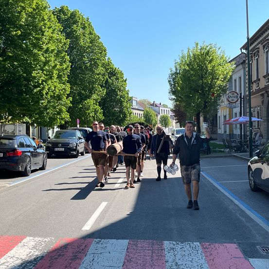 Eine Gruppe von Menschen in Kilts und mit Holzfässern geht eine Straße entlang, an der Autos auf beiden Seiten geparkt sind.