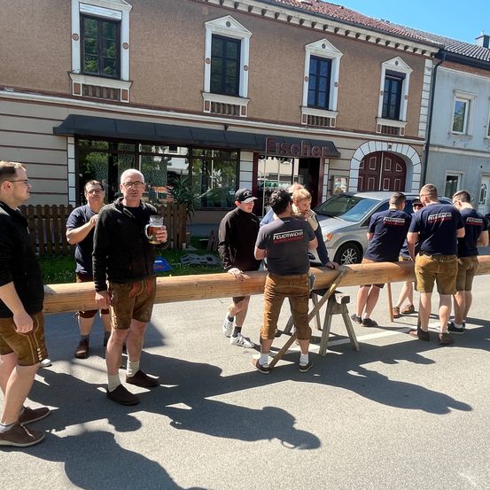 Eine Gruppe von Männern in traditioneller deutscher Kleidung trägt einen großen Baumstamm vor einem Gebäude mit dem Schild 'Fischer'.