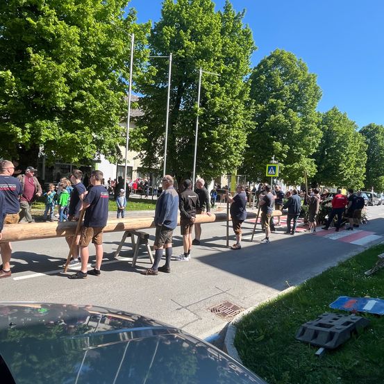 Mehrere Menschen sind auf der Straße und tragen einen langen Holzbalken. Ein Straßenschild steht am Straßenrand.