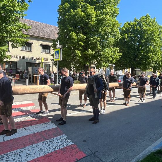 Eine Gruppe von Männern, die einen langen Holzbalken über eine Straße vor einem Gebäude mit vielen Menschen im Hintergrund tragen.