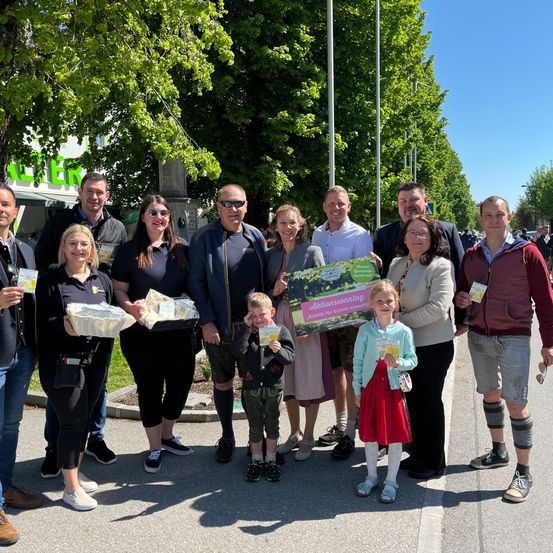 Eine Gruppe von Menschen steht vor einem Baum. Sie halten ein grünes Plakat, eine Box und ein Büchlein. Die Menschen tragen Freizeitkleidung.