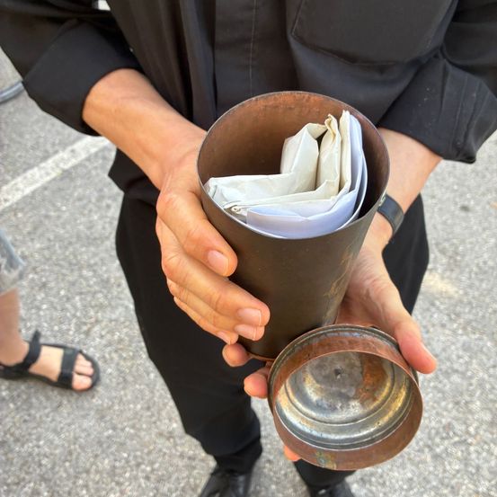 Eine Person in Schwarz hält einen Kupferbehälter mit gefalteten Papieren im Inneren, steht im Freien mit einer weiteren Person in Sandalen.