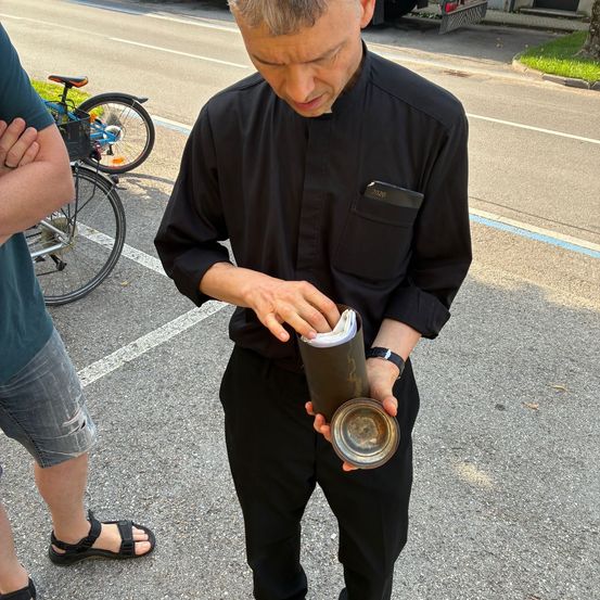 Ein Mann in schwarzer Kleidung, möglicherweise ein Priester, steht im Freien mit gefalteter Papier in den Händen, in der Nähe eines Fahrrads und einer weiteren Person.