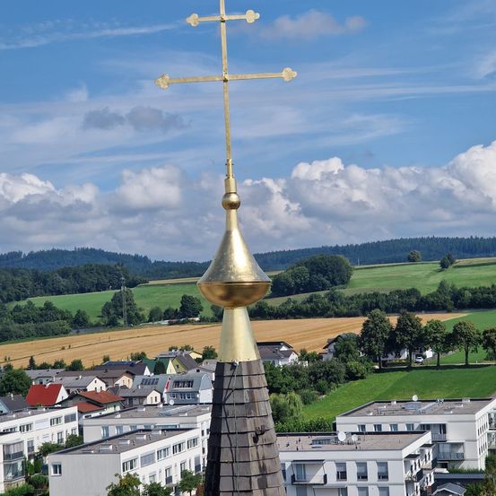 Ein Blick von einem Turm auf ein goldenes Kreuz oben mit einem Dorf darunter, grünen Feldern und einer Bergkette in der Ferne.