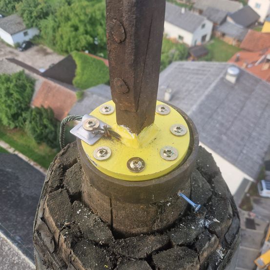 Ein Nahaufnahme eines Stahlmastes mit gelber Basis, mehreren Schrauben und einem Bolzen, der an einer hölzernen Struktur befestigt ist, von oben über einer Vorstadt mit Häusern und Grünflächen betrachtet.