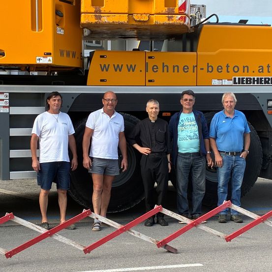 Fünf Männer posieren vor einem gelben Liebherr-Kran. Die Männer tragen lässige Sommerkleidung. Zwei tragen eine Brille und einer eine Halskette. Der Boden ist mit grauem Beton gepflastert.